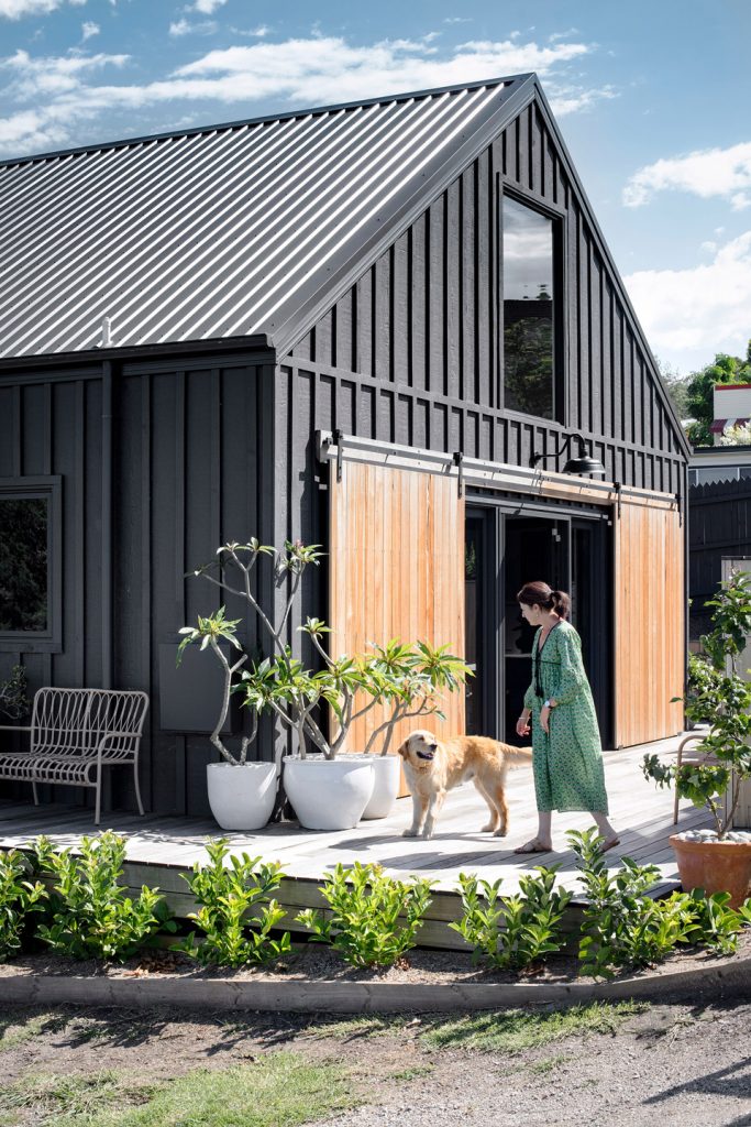 A lady and her dog standing in front of a barn style home built in Weathergroove woodsman 1200 battens are used painted in black with the doors in a wood finish.