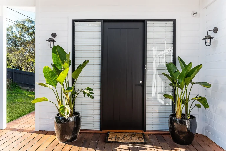 Front of white house with black cladded boards single door. Two pot plants sit either side